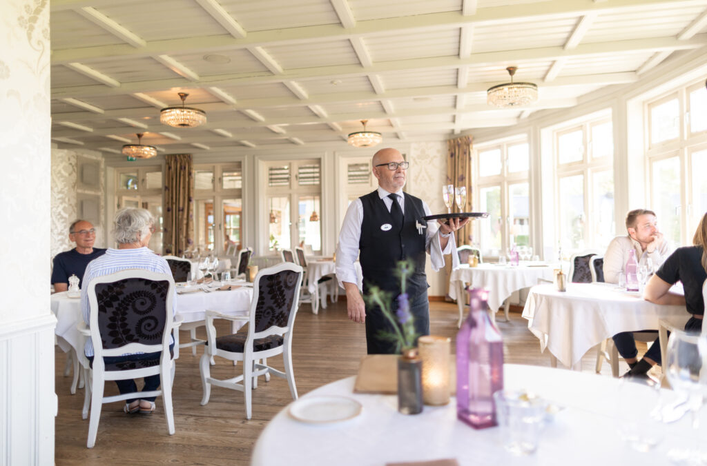 Vy över en av matsalarna på Hestraviken med servitör och gäster vid bord.