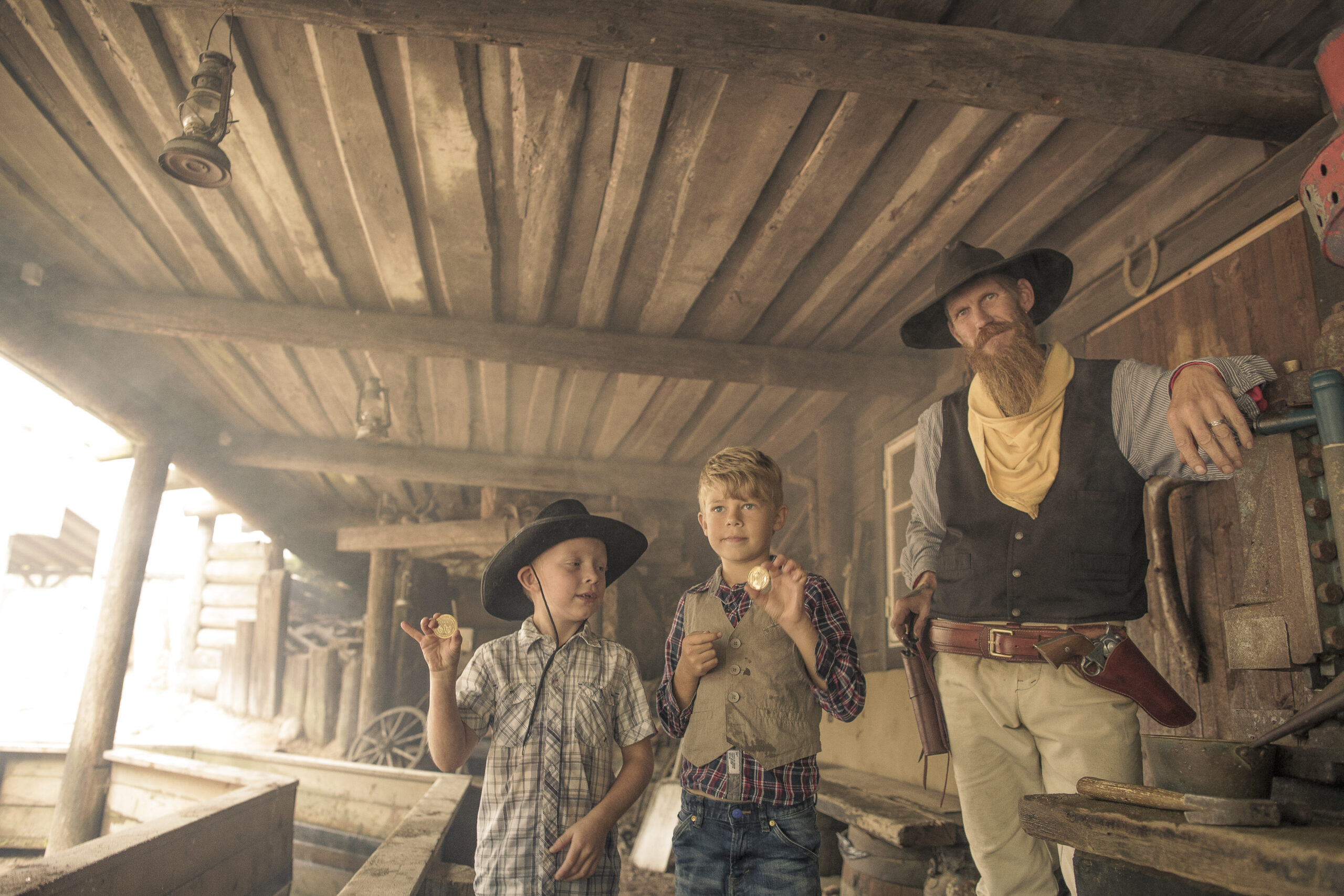 Två barn klädda i rutiga skjortor och med cowboyhatt tillsammans med en cowboy i väst, hatt och gul scarf som vaskar fram guld ur en ränna av trä, av guldet tillverkas ett mynt som barnen håller upp.